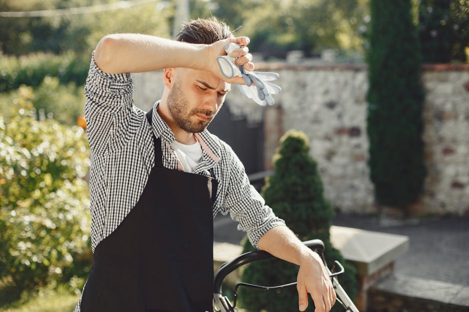 A man stands outdoors in a garden on a sunny day, wearing a black apron over a checkered shirt, holding a pair of garden gloves in his right hand, which he is resting on his forehead. The garden featu